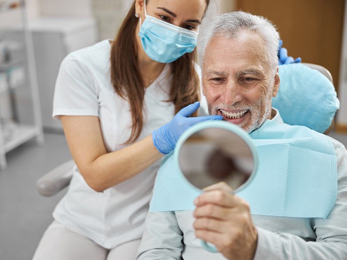 Man smiling into handheld mirror with masked dentist touching his teeth